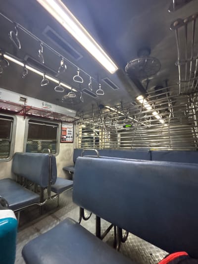 The image depicts the interior of a commuter train, likely in or near Ambernath, India. Visible are rows of blue padded seats, metal overhead bars and handholds, ventilation fans, and fluorescent lighting. A poster is visible on the wall. The scene is typical of a public transportation setting, devoid of passengers. The perspective is from within the train car, looking towards the end.