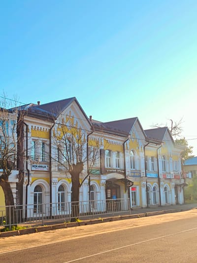 The image depicts a street view in the city of Kimry, Russia. A multi-story building with a yellow and white facade stands prominently on the right side of the frame. The building features several arched windows and ornate architectural details. A dark-colored metal roof caps the structure. Bare trees with intricate branches are situated in front of the building, suggesting a cooler season. A metal fence runs along the sidewalk, separating it from the street. The street itself is paved and appears to be a road with visible lane markings. Sunlight casts long shadows, indicating it is either early morning or late afternoon. The sky is clear and a bright blue. Several signs with Cyrillic text are visible on the building's facade, indicating shops or businesses. No people are clearly visible in the image. The overall scene is one of an urban environment with a focus on the architectural character of the building.