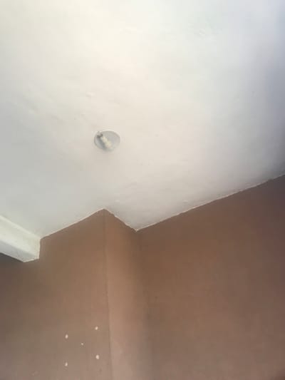 The image captures an interior architectural corner, primarily featuring two intersecting surfaces painted in contrasting colors: white and brown/tan. The dominant feature is a white wall, occupying the left and central parts of the frame. Mounted on this white wall, slightly below the center, is a simple, circular light fixture, likely a bulb holder, with a metallic component. The white wall exhibits some textural variations and minor unevenness in its paint finish.
To the right and top of the white wall, a brown or tan-colored surface extends, forming a distinct corner. This brown surface appears to be a ceiling or an upper wall segment. It also shows textural qualities and contains a few small, scattered white specks, possibly paint splatters or debris, particularly in the lower right portion of its area.
In the bottom right corner of the image, an additional white, rectangular structural element or trim is visible, indicating another architectural junction. The lighting is even, suggesting an indoor setting. There are no people or discernible activities taking place, and no visible text within the frame. The architectural elements presented are generic and do not offer specific visual cues that uniquely identify the stated location of Umudike, Nigeria.