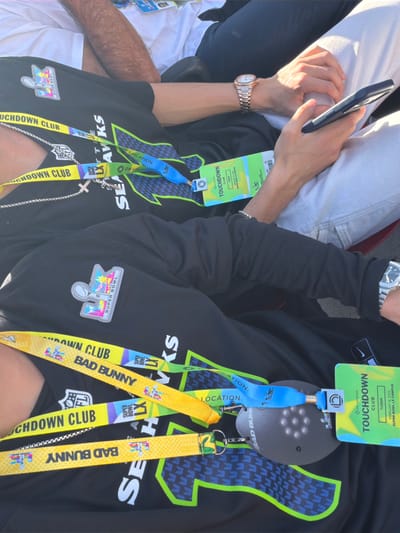 Two people wearing Seattle Seahawks apparel and Super Bowl badges are seated close together. The badges indicate access to the Touchdown Club and feature a yellow Bad Bunny lanyard. One person is holding a phone and wearing a watch. Both individuals have Super Bowl LX patches on their clothing. The badges also have an On Location logo and list the date as February 8, 2026. The overall scene suggests they are attending the Super Bowl LX campus.