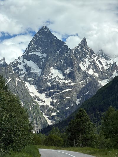 A wide-angle, eye-level, outdoor shot captures a majestic mountain range under a cloudy sky. The dominant feature is a massive, jagged mountain peak, partially covered in snow, with steep rocky slopes. To its right, a smaller, sharp peak pierces the clouds. The lower slopes of the mountains are covered in lush green trees, forming a dense forest. In the foreground, a paved road with white lane markings curves from the bottom left towards the right, bordered by grassy verges. The scene is tranquil and untouched, suggesting a remote, natural setting. The location is identified as Верхне-Тебердинское сельское поселение, Russia. There are no people or animals visible in the image.