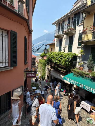 A street scene in San Giovanni, Italy, features people strolling along a cobblestone path lined with buildings. The buildings have colorful facades and architectural details like shutters and balconies. One building displays a green awning with the text "'DA GANCIO' TRATTORIA" and "APERITIVI WINE BAR," suggesting a restaurant or bar is present. People are dressed in casual summer attire, indicating a warm, sunny day. Some individuals are seated at tables under the awning, while others are walking or standing, engaged in conversation or looking at their surroundings. A few promotional signs are visible, including one that says "WINE KIT TO GO." The overall atmosphere appears relaxed and social, typical of a pleasant day in a European town. The presence of a lake and mountains in the background suggests a scenic, possibly coastal or lakeside, location. The time of day seems to be late morning or afternoon, given the sunlight and shadows.