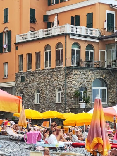 The image depicts a crowded beach scene in Camogli, Italy, with a multi-story building serving as a backdrop. The building features an orange facade with various windows and balconies, some with green shutters. A stone-clad lower section of the building is visible, punctuated by arched windows. In the foreground, numerous beach umbrellas, mostly yellow and orange, provide shade for beachgoers. People are seen lounging on beach chairs, some in swimwear, and others appear to be relaxing or socializing. The beach itself is covered in grey pebbles. A date and time stamp, "31.07.2026 18:27", is present in the lower right corner. The overall atmosphere is one of a sunny day at a popular seaside resort.