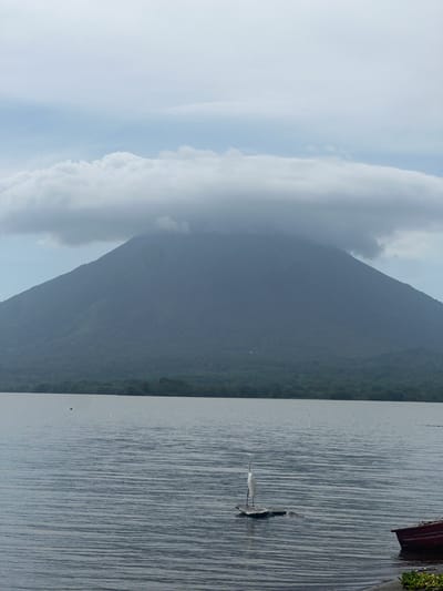 The image captures a serene landscape in Altagracia, Nicaragua, featuring the imposing Mombacho volcano partially obscured by thick, white clouds. The volcano rises majestically in the background, its slopes covered in lush, dark green vegetation. In the foreground, a calm body of water reflects the overcast sky, with gentle ripples disturbing the surface. A lone white heron stands gracefully on a small, dark object in the water, appearing as a solitary figure in the vast expanse. To the right, the corner of a red boat is visible at the water's edge, hinting at human presence and activity in the area. The overall atmosphere is tranquil and slightly melancholic due to the cloudy weather.