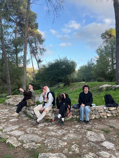 Four individuals are depicted resting on a low, weathered stone wall in an outdoor, semi-forested environment within the Megido Regional Council, Israel. The ground in the foreground is paved with irregular flat stones, leading to dirt and sparse green vegetation beyond the wall. Tall, slender trees, primarily conifers, rise on both sides of the frame and in the background, under a blue sky with scattered white cumulus clouds.

From left to right:
1.  A male with short dark hair, wearing a dark brown jacket over a grey shirt and dark trousers, sits on the wall. His right arm is raised, with his hand positioned as if holding a device or gesturing towards the left. A light-colored shoulder bag is visible across his body.
2.  A male with a red bucket hat and a light grey hooded sweatshirt, light ripped trousers, and dark shoes, sits next to the first individual. A black backpack with a yellow water bottle attached hangs from his left shoulder. He holds a lit cigarette or similar item to his mouth with his right hand, exhaling. A clear plastic bottle with a red label rests on the ground in front of him.
3.  A person with dark hair, wearing a black hooded top and dark trousers, is seated with their head down, appearing to rest or sleep, slumped forward over their knees. A red and black plaid garment is visible around their waist or lap. A small black bag is placed on the wall next to them.
4.  A male with curly dark hair, wearing a black hooded sweatshirt and light blue jeans, sits upright. His hands are clasped in front of him, and he gazes towards the right. A black backpack rests on the ground to his right, and a white container is on the wall beside him.

The environment suggests a natural area, possibly a park or trail, with a dirt path visible behind the group, leading into denser vegetation and rolling hills. The lighting indicates daylight conditions.