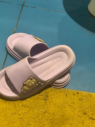 Two light purple slides with a cartoon bear design are placed on a textured blue surface, with a yellow mat partially visible in the foreground. The slides are made of a smooth material, possibly rubber or plastic, and feature a thick sole with ridged detailing. One slide is positioned in front of the other, overlapping slightly. The cartoon bear decoration on the front strap of each slide is light brown with darker brown accents for its eyes and nose. The blue surface appears to be a tiled floor, possibly in a bathroom or a spa area, with thin grout lines visible. The yellow mat is fluffy and textured. The lighting suggests it could be daytime, with some shadows indicating light coming from the side. There is no text visible in the image. The overall scene is casual and domestic.
