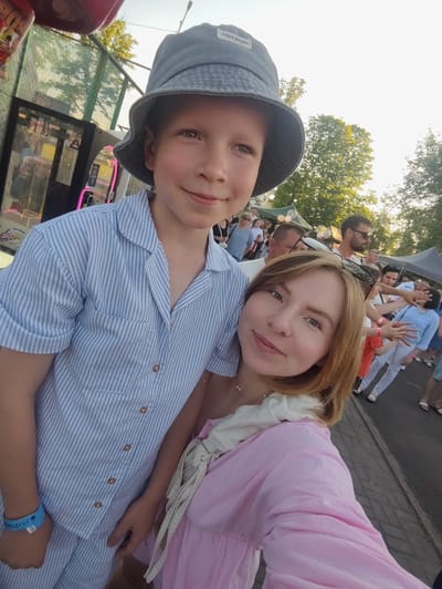 This is a selfie taken outdoors in Korosten, Ukraine, likely at an amusement park or fair. A young boy wearing a blue and white striped shirt and a grey bucket hat is in the foreground, looking towards the camera with a smile. To his right, a woman with blonde hair is also looking at the camera. She is wearing a pink dress and a white scarf. In the background, there are several other people, trees, and structures, including what appears to be an arcade game machine and tents. The sun is setting, casting a warm light on the scene.