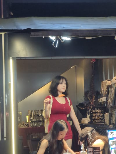 In Longta, China, at night, a young woman in a red dress is seen holding a skewer with food. She has dark hair and is standing in front of a stall selling jewelry. To her left, a vertical fluorescent light illuminates the area. In the foreground, another young woman, partially visible, is looking down. The background shows shelves filled with various necklaces and earrings. The scene appears to be outdoors, possibly a market or street vendor area.
