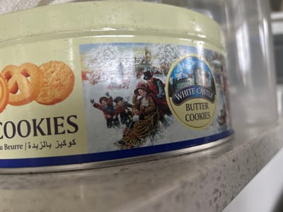 A close-up, eye-level shot shows a round, light-yellow metal tin of "WHITE CASTLE BUTTER COOKIES" resting on a speckled grey countertop. The tin features a Christmas-themed illustration depicting figures in period clothing sledding down a snow-covered hill with a large building in the background. The left side of the tin displays images of three butter cookies, two of which are ring-shaped and one is round. Text on the tin includes "COOKIES," "Au Beurre," and Arabic script. The background is blurred, with a clear plastic container to the right and what appears to be a white textured surface above. The scene is indoors, likely in a kitchen or pantry. The location of Richmond Hill, Canada is not discernable from the image.