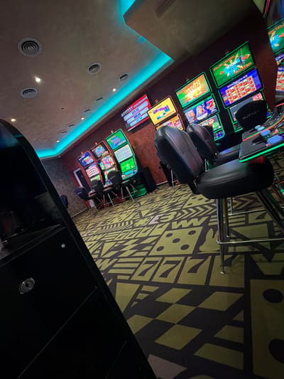 The image captures the interior of what appears to be a casino, possibly located in Graf Ignatievo, Bulgaria. Rows of slot machines with illuminated screens displaying different game titles are visible along the wall on the left. Swiveling chairs are positioned in front of the machines for patrons to sit on while playing. The floor is covered with patterned carpeting. The ceiling has recessed lights, and the walls seem to have ambient lighting. A black counter or bar is present in the foreground.