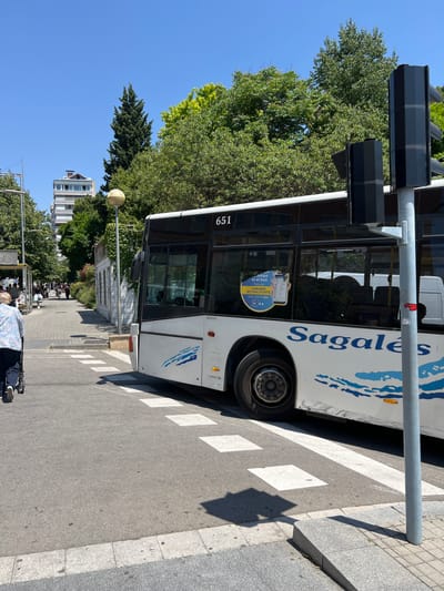 The image captures a bus of the Sagalés company at a bus stop located on a street with a crosswalk. The bus, identified by the number 651, is white with blue markings and has promotional materials about the bus schedule and app displayed on its windows. A traffic light post is visible, and trees line the street. In the background, a building can be seen. An elderly person with a caretaker can be seen waiting at the bus stop. The location is likely in Granollers, Spain, as Sagalés is a bus company that operates in the region.