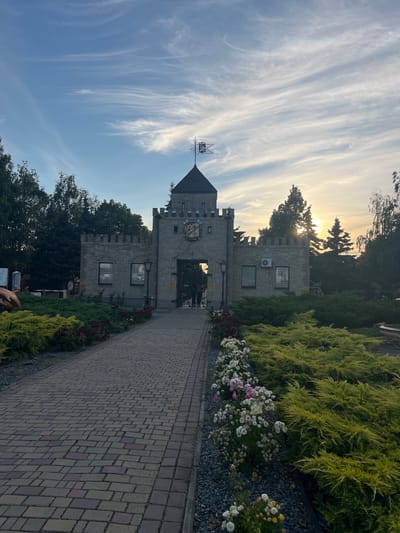 This is a vertical, low-angle shot of a whimsical, castle-like structure on a sunny day. The building is made of light-colored stone blocks and features battlements, arched doorways, and several windows. A weather vane with a detailed metallic design is visible atop one of the towers.

The scene is framed by lush green foliage and a row of blooming flowers in shades of white and pink. A paved pathway made of rectangular stones leads towards the entrance of the structure. The sky above is a clear blue, streaked with wispy white clouds, suggesting fair weather. The sun is low in the sky, casting a warm glow and creating a gentle lens flare as it peeks through the trees, indicating it is either sunrise or sunset.

There is some visible text on yellow rectangular signs affixed to the building and on what appears to be a traffic barrier. One sign near the entrance has red lettering, possibly indicating "Stop" or a prohibition. Another sign on the building has text that is too small to read.

The overall impression is one of a charming, possibly themed, destination. The soft lighting and vibrant greenery create a peaceful and inviting atmosphere. The location appears to be in Slobozhanske, Ukraine.
