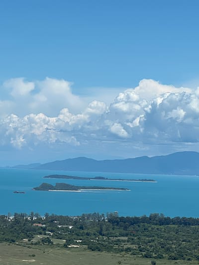 An aerial view captures the turquoise waters of the Gulf of Thailand, dotted with lush green islands and a distant mountain range under a bright blue sky filled with fluffy white clouds. In the foreground, a verdant landscape unfolds, featuring a dense forest interspersed with scattered buildings, suggesting a settlement within Baan Thurian, Thailand. The scene evokes a sense of tropical tranquility.