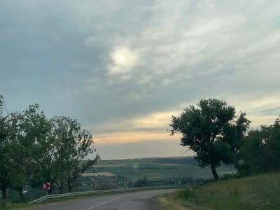 The image depicts a scenic view of a rural landscape in Albota de Jos, Moldova. The scene is dominated by a winding road that curves into the distance, flanked by green trees and lush vegetation. In the foreground, two trees with dense foliage stand prominently on either side of the road. The road itself is paved and appears to be a two-lane street. Road signs with red and white markings indicate curves in the road.
Beyond the road, the landscape unfolds into rolling hills covered in a patchwork of fields and scattered houses, suggesting a small village or settlement. The sky is overcast with dramatic clouds, but a bright spot of sunlight pierces through, illuminating the scene with a soft, diffused light. The time of day appears to be late afternoon or early evening, given the muted sunlight and the overall atmospheric quality. The weather seems to be calm, with no visible signs of rain.
The overall impression is one of tranquility and natural beauty, characteristic of a peaceful countryside setting. There is no visible text in the image.
