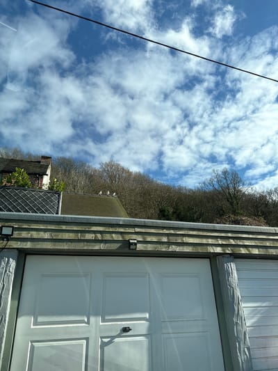 The image shows a view in Snailbeach, United Kingdom. A white door with rectangular panels and a black handle is visible on the right. To the left of the door is a grey, vertically slatted wooden wall with two square light fixtures. Beyond the wall are other buildings, some with visible brickwork and dark-colored roofs. Above the buildings are bare trees against a bright blue sky with scattered white clouds. A black powerline cuts diagonally across the upper left corner of the image. Some white birds can be seen perched on the roof of a building.