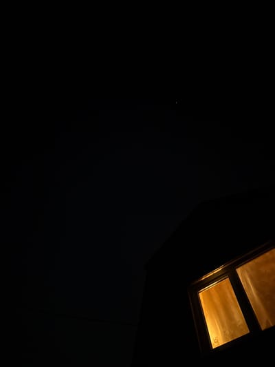 A dark, vertical, full shot image shows a night sky with a few faint stars. The bottom right corner of the image is dominated by the silhouette of a dark structure, likely a building. A window within this structure is illuminated from within by a warm, orange light, suggesting the presence of an interior light source. The window is divided into two panes, both covered by sheer, translucent curtains. The curtains are a light yellow or beige color and show faint patterns from the light behind them. The overall scene conveys a sense of quiet and domesticity at night. The location is not specified in the image itself, therefore the reference to Ermakovo, Russia is an external detail.