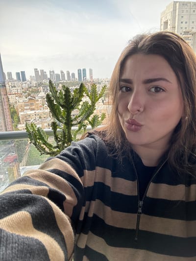A female subject is depicted in a selfie from a high-rise balcony in Bnei Brak, Israel, with a westward view. She possesses shoulder-length brown hair, a nose ring on her left nostril, and wears a black and beige striped quarter-zip sweater. Her lips are pursed in a pout. Behind a grey metal balcony railing, a multi-stemmed green cactus-like plant is present. The background displays a dense urban landscape consisting of residential buildings in Bnei Brak, leading to a prominent cluster of modern high-rise buildings characteristic of the Tel Aviv skyline in the mid-distance, all under an overcast sky.