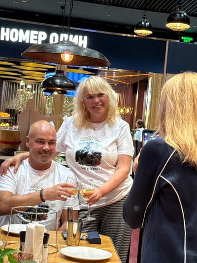 The image shows three individuals in what appears to be a restaurant or establishment with signage in Russian that translates to "NUMBER ONE" and "EXIT." A man and a woman, both wearing white t-shirts, are in the foreground. The man, with a shaved head, is smiling and has his arm around the woman's shoulders. The woman, with blonde hair, is also smiling. She is wearing a t-shirt with a graphic of a mountain and text in Russian. A third person, partially obscured from the back and with blonde hair, is holding a smartphone and taking a picture of the couple. The background shows decorative lighting fixtures and a dimly lit interior. The scene is set on a table with plates, glasses, and cutlery. The location is identified as Perepechino, Russia.
