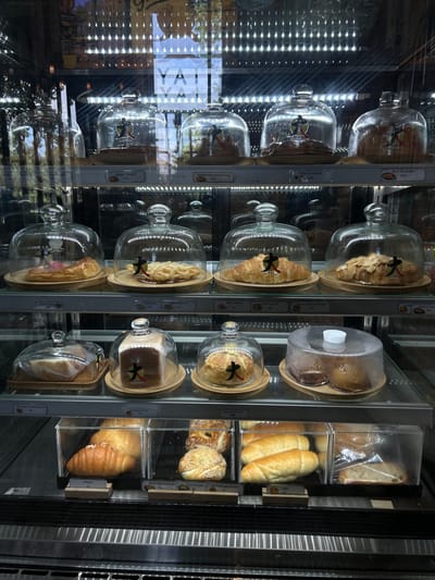 The image shows a refrigerated display case in a bakery in Prachuap Khiri Khan, Thailand, filled with various baked goods. The display case has multiple shelves, each illuminated by overhead lights. The baked goods are primarily placed on wooden bases and covered with glass domes or plastic wrap.

On the top shelf, several items are visible, including what appear to be danishes and croissants, some with toppings like nuts. Labels with prices and item names in Thai and English are placed below some of these items.

The second shelf features a variety of pastries, including croissants, a slice of cake or tart, and what looks like a waffle or pastry with a criss-cross pattern. Again, labels are present for some items.

The third shelf contains loaves of bread, some sliced, and smaller buns or pastries. One item is a white loaf of bread covered by a glass dome, and another is a darker loaf covered by plastic wrap.

The bottom shelf displays more bread rolls, croissants, and some round, sesame-seed topped buns. These are mostly contained in clear plastic boxes.

The overall environment suggests a commercial bakery or cafe setting. The lighting is bright, reflecting off the glass surfaces of the display case. There are no people visible in the image, and the focus is entirely on the displayed food items. The branding on the baked goods appears to be consistent, featuring a stylized logo.