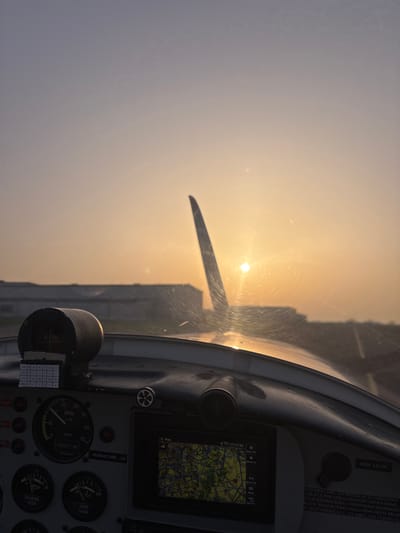 The scene depicts the interior of a light, single-engine aircraft cockpit, viewed through a dirty or scratched windshield, during either sunrise or sunset. The sun is low on the horizon, appearing as a bright, circular disc slightly right of center, casting a warm orange glow across the hazy sky and causing significant glare on the windshield. A single propeller blade is silhouetted against the sun. In the background, a large, dark building (likely a hangar) and a paved airfield surface are visible. The aircraft's instrument panel occupies the lower portion of the image, displaying analog gauges for speed (in km/h, indicating near zero), oil pressure, oil temperature, and fuel reserve. Centrally positioned on the panel is a digital navigation screen showing a map of the Toussus-le-Noble, France area, consistent with Toussus-le-Noble Airport (LFPN), and displaying a time of "17:12:44". No individuals are visible. The aircraft appears to be stationary or taxiing slowly on the ground at Toussus-le-Noble Airport in Toussus-le-Noble, France.
