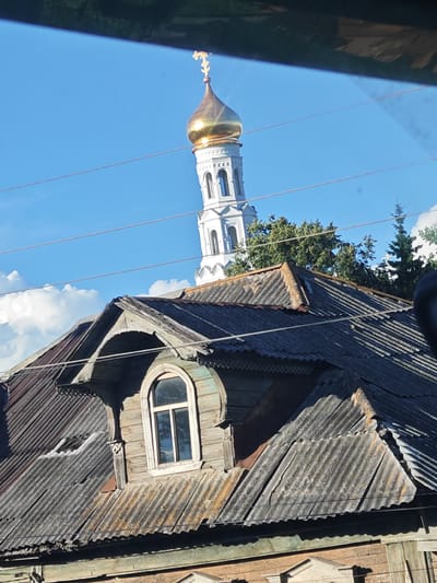 In Zavidovo, Russia, the image captures a view of a weathered, old wooden house with a corrugated metal roof in the foreground. The roof has visible signs of age and damage. Above the house, the golden dome of a white church tower, adorned with a cross, stands against a clear blue sky with scattered white clouds. Several utility wires stretch across the frame, adding to the urban or semi-urban setting. Trees, including coniferous ones, are visible behind the house and to the right of the church tower. The overall scene suggests a historical or traditional architectural style within a Russian town or village.