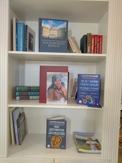 The image shows a close-up view of a white, multi-shelf bookcase filled with books. The bookcase is positioned within a room with white, vertically fluted decorative elements on the right side, suggesting it is part of a larger piece of furniture or a built-in. The lighting appears to be artificial and evenly distributed, with no discernible natural light sources. The top shelf contains several books. On the far left, a book with a spine that appears to be blue and white, with text in Russian, is partially visible. To its right, a larger, dark blue book prominently displays the title "ИМПЕРАТОРСКИЕ РЕЗИДЕНЦИИ МОСКОВСКОГО КРЕМЛЯ" (Imperial Residences of the Moscow Kremlin) in large, gold-colored Russian lettering, accompanied by an image of a grand building. Several other books with visible Russian titles are also on this shelf, with spines in various colors including red, brown, and green. The middle shelf features a framed portrait of a young girl wearing a blue headscarf and pearl necklace, with a warm smile. The frame is a deep red color, and text in Russian is visible along its side. To the left of the portrait, a stack of three books is arranged vertically. The top book has a green spine with gold lettering. The two books below it have blue and white patterned spines, with Russian text, including titles like "ГОСУДАРЬ ИМПЕРАТОР" (Sovereign Emperor). To the right of the portrait, a single book with a dark blue cover is titled "АЛЕКСЕЙ МОСКАЛЕВ" (Alexey Moskalev) and "120 ЛЕТ ЖИЗНИ. ТОЛЬКО НАЧАЛО." (120 Years of Life. Only the Beginning.) in large white text. The subtitle reads "КАК ПОБЕДИТЬ СТАРЕНИЕ?" (How to Defeat Aging?). This book also contains graphics of gears and text in Russian. The bottom shelf contains several books. On the left, a stack of three books are leaning against each other. The foremost book on the right is a large book with a dark blue cover, titled "АЛЕКСЕЙ МОСКАЛЕВ" (Alexey Moskalev) and "МОЗГ ДОЛГОЖИТЕЛЯ" (Brain of a Long-Lived Person). It features text in Russian and an illustration of a head with gears inside. To the left of this book, three other books are partially visible, with spines displaying Russian text and patterns. One of these books has the title "ФЕДОТ ВАСИЛЬЕВИЧ СЫЧКОВ" (Fedot Vasilyevich Sychkov). On the right side of this shelf, a book with a colorful cover depicting a woman in traditional clothing is open,