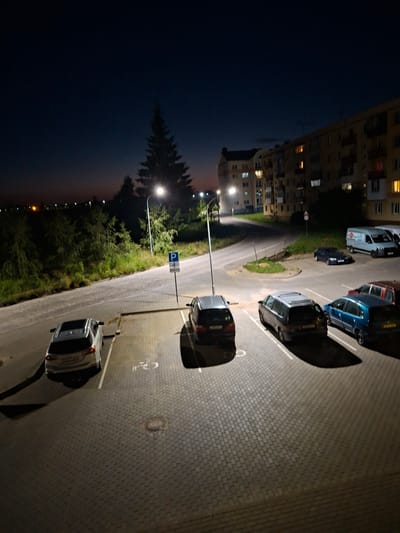 A nighttime, medium-height, wide-angle view captures a residential complex and a parking lot in Dzyarzhynsk, Belarus. The sky is dark blue, transitioning to a faint orange glow on the horizon. In the background, a multi-story apartment building with lit windows stands against the night sky. A tall evergreen tree is illuminated by a street lamp. The foreground features a paved parking lot with several parked cars, including a silver SUV, a dark grey minivan, and a blue station wagon. The parking lot is bordered by a sidewalk and a grassy area with more trees. The overall scene is dimly lit by artificial lights, creating long shadows.