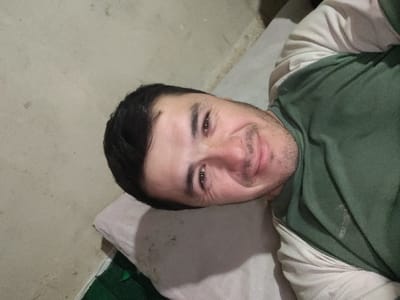 An adult male is depicted in an indoor setting, identified as the city of Yangikurgan, Uzbekistan. He is lying down on his back, supported by a light-colored pillow, with his head turned slightly towards the viewer. The man has short dark hair, brown eyes, and stubble on his face, and is presenting a faint smile with visible wrinkles around his eyes. He is wearing a two-toned shirt, featuring a dark green upper layer over a lighter-colored, possibly beige or off-white, long-sleeved undershirt. A portion of a dark green mat or textile is visible on the floor to the left of the pillow. The background consists of a plain, light-colored wall, possibly beige or off-white, exhibiting minor surface imperfections. The scene is evenly lit, indicating an interior environment.