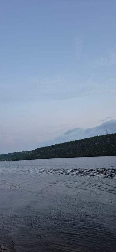 A wide, dark body of water occupies the foreground and midground of the image, with gentle ripples indicating calm movement. In the background, a densely vegetated, hilly landscape stretches across the horizon. The vegetation is predominantly dark green, suggesting trees or shrubs. On the crest of the hill, a lone, tall transmission tower with a lattice structure is visible on the right side. The sky above is a pale blue, with some thin, wispy clouds scattered across it, particularly towards the horizon where they appear to be denser and darker. The overall lighting suggests it is either early morning or late evening. The scene appears to be a natural landscape, possibly a river or a large lake, with the hilly terrain on the opposite bank. The location is the city of Molovata Nouă, Moldova.