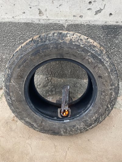 An overhead view shows a worn tire lying flat on a sandy surface in front of a textured concrete wall. A small metal contraption, with a flickering flame visible inside, is positioned within the center of the tire. The tire is dirty and shows signs of use. This scene is located in Kwalla, Nigeria.