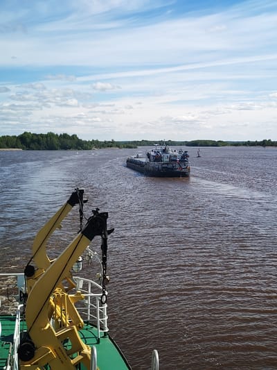 The image shows a river scene in what appears to be Kargulino, Russia. A river barge is mid-stream, heading away from the camera. The riverbanks are lined with trees. The sky is partly cloudy. In the foreground, the yellow and black cranes of a ship are visible, partially obscuring the view. The water is dark brown.