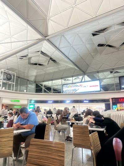 The image depicts an interior scene at Stansted Airport in Uttlesford, United Kingdom. The architecture features a distinctive geometric ceiling with white panels and metal supports. In the foreground, several individuals are seated at tables, some engaged with electronic devices, others conversing, and one person is asleep. Various individuals are visible in the background, indicating a busy public space. Signage for "Health," "Beauty," and a "Coffee" establishment are discernible. A large digital billboard displays an advertisement with the text "NO DRAMA" and "GOING TO CENTRAL LONDON IS FASTER BY TRAIN." A large industrial fan is mounted on a metal structure to the left. The overall environment appears to be a food court or seating area within an airport terminal.
