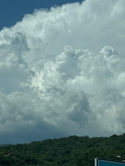 The image shows a dramatic sky filled with large, white cumulus clouds against a bright blue sky. The clouds have a sense of depth and texture, with areas of light and shadow suggesting their immense size and form. Below the clouds, a lush, green hillside slopes upwards, densely covered in trees. The terrain appears to be somewhat rugged, with winding lines visible within the foliage, possibly indicating paths or terraces. On the hillside, a small, light-colored building or structure is partially visible, nestled among the trees on the left side of the frame. The very bottom right corner of the image shows a portion of a blue and white sign. The overall scene suggests a natural landscape, possibly in a mountainous or hilly region, under a dynamic sky. The specific location of Vrijesno, Montenegro is not depicted visually within the image itself, but is provided as context for the scene.