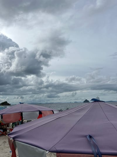 The image depicts a beach scene with several beach umbrellas open under a cloudy sky. The umbrellas are in various colors, including purple, pink, and white. Some people are visible beneath the umbrellas, suggesting they are enjoying a day at the beach. The sea is visible in the background, with a distant island or landmass on the horizon. The sky is filled with dramatic, dark clouds, indicating overcast or possibly stormy weather. The overall impression is that of a beach day interrupted or characterized by inclement weather, yet people are still present and utilizing the beach amenities. The location is identified as Tambon Phru Nai, Thailand, which is a coastal area. No text is visible in the image. The lighting suggests it is daytime, but the heavy cloud cover obscures direct sunlight.