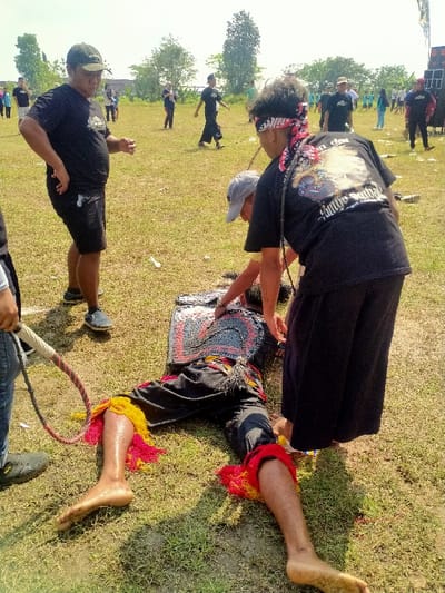 This image depicts an outdoor scene in Celep, Indonesia, under daylight conditions. A person lies supine on a grassy field, barefoot, dressed in dark pants with prominent red and yellow fringed decorations at the ankles and a multi-colored, patterned chest covering. Two individuals are positioned over the person on the ground: one, wearing a grey baseball cap and dark t-shirt, leans forward towards the chest area; the other, wearing a dark t-shirt with a graphic on the back, dark wide-legged pants, and a red and white patterned bandana around their head, also leans over the prone individual. A dark braided rope or whip is visible near the second individual. To the left, a man in a dark t-shirt, dark shorts, and a baseball cap stands observing, with his hands on his hips. In the far left foreground, the lower body of another individual is visible, holding a whip with a braided handle. The background features an expansive grassy area with several individuals scattered throughout, mostly dressed in dark casual clothing. Trees line the horizon, and a large black sound system is visible on the far right. The ground consists of green grass with exposed patches of dirt.