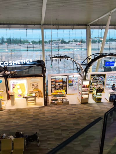 The image shows an interior view of a duty-free shop in Tambon Mai Khao, Thailand, likely at an airport. The shop is well-lit and displays a variety of cosmetic products on shelves and stands. Prominently featured is a display for Chloe perfume and other beauty items. In the background, large windows offer a view of an airport tarmac with airplanes parked and taxiing, indicating the location is an airport terminal. The flooring is carpeted with a patterned design. In the foreground, a person is seated in a wheelchair next to some bags, and two chairs are visible. The overall scene suggests a commercial area within a transportation hub.