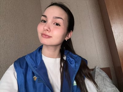 An image taken indoors in what is identified as the city of Bayronovka, Russia, depicts a young adult female from the chest up, positioned slightly off-center and looking directly at the camera. She has light skin, dark eyes, and long dark hair, parted on her left and largely falling behind her shoulders, with some strands visible on her right. She exhibits a subtle smile with closed lips. Her right ear features a small, silver-colored hoop earring with a clear stone. She is dressed in a white t-shirt topped with a blue vest. The vest has a yellow square logo on the left chest area, accompanied by partially visible Cyrillic text that appears to reference a "Presidential Grants Foundation." The background consists of a light beige or light brown textured wall on her left and a dark brown wooden furniture panel on her right. A portion of a bed with a pillow featuring a grey and white botanical pattern is visible in the lower right background.