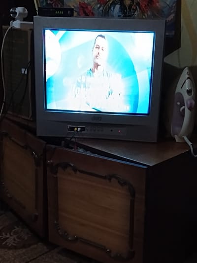 A television is on a wooden cabinet in Luhansk, Ukraine. The television is displaying a man with short dark hair and a light-colored shirt. To the left of the television, a white webcam is visible. To the right of the television, a white and pink iron is on the cabinet. The television is connected to a black box with red and yellow wires. The wooden cabinet has ornate metal decorations on its doors. The room appears to be dimly lit, with a patterned rug visible on the floor.