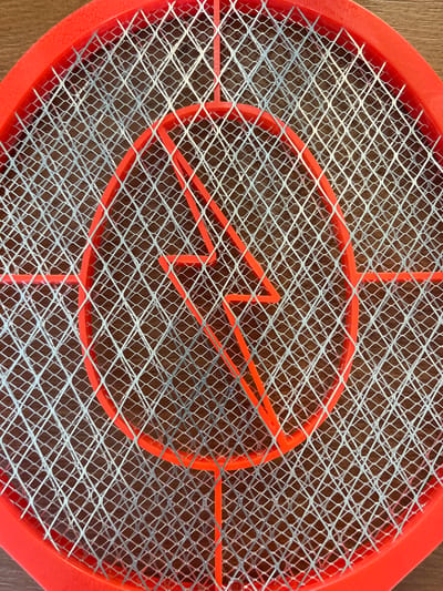 The image is a close-up, overhead shot of an electric mosquito swatter. The swatter has a bright orange plastic frame and a diamond-patterned metal mesh grid. Centered within the grid is an orange plastic emblem shaped like a lightning bolt within a circle. The swatter rests on a wooden surface, likely a table or floor, with a visible wood grain pattern. The lighting appears to be artificial, casting subtle shadows on the wood. The scene is static and devoid of any activity. The location of the image, in Lastva Grbaljska, Montenegro, is not visually represented in the photograph itself; it is external information provided with the image.