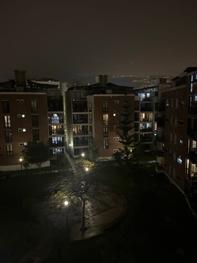 The image depicts a courtyard surrounded by multi-story brick buildings in Santiago de Surco, Peru, at night. The sky is overcast. The buildings have lighted windows. A tree with lights strung through its branches stands in the courtyard. There is a partially visible semicircular structure behind the tree.