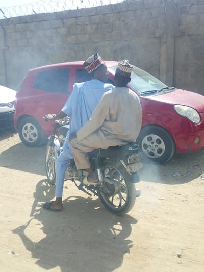 The image depicts two adult males on a motorcycle, stationary or moving slowly, on a dirt path in Darmanawa, Nigeria. The driver, wearing a light blue traditional garment and a patterned cap, is positioned on the front seat. The passenger, wearing a light tan traditional garment and a different patterned cap, sits behind the driver. Both individuals are facing forward, with the passenger's head turned slightly towards the driver. The motorcycle is dark-colored with chrome elements, and white exhaust smoke emanates from its rear.
In the background, a red two-door hatchback car is parked to the right of the motorcycle, facing left, partially obscured by the motorcycle and its occupants. A portion of a white vehicle is visible to the far left. Behind the vehicles, a weathered concrete wall topped with coiled barbed wire runs horizontally across the upper third of the frame. The ground is unpaved, consisting of compacted dirt with visible tire tracks. Strong sunlight from a high angle casts distinct shadows of the motorcycle and its occupants onto the ground, directly below and to the left. The motorcycle seat features the brand name "Daylong."