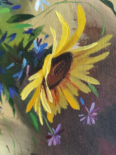 The image is a close-up, angled photograph of an oil painting. The painting depicts a sunflower in the foreground, with its bright yellow petals dominating the center of the frame. The sunflower's center is a dark brown, textured circle. To the left of the sunflower, there is a bouquet of smaller flowers, rendered in shades of blue and white, interspersed with green leaves. The background is a textured, dark brown. In the lower right quadrant of the painting, there are small, purple flowers with delicate petals. The overall style of the painting is impressionistic, with visible brushstrokes and a vibrant color palette. The image does not contain any information or visual cues that would allow for the identification of its location within the city of Rybnitsa, Moldova.