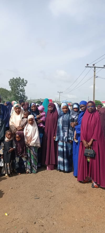 A group of approximately fifteen people, primarily women and children, are standing together outdoors. Most of the women are wearing hijabs and long, flowing gowns of various colors, including maroon, blue, beige, and patterned fabrics. Some are holding babies or young children. The children in the foreground are wearing similar styles of clothing.
The setting appears to be a dusty, open area with a few patches of vegetation. In the background, there is a utility pole with wires and a scattering of buildings, suggesting an urban or semi-urban environment. The overcast sky suggests a daytime scene. The location is Naraguta Hausawa, Nigeria. The group is stationary and appears to be posing for a photograph.