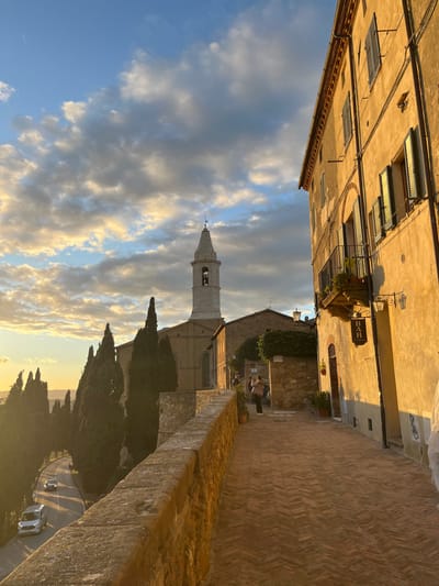 This image depicts a picturesque street scene in Pienza, Italy, bathed in the warm, golden light of late afternoon or early evening. The overall atmosphere is tranquil and inviting.

In the foreground, a narrow, paved street or alleyway, possibly made of brick, leads into the distance. To the left, an old, light-colored building rises, featuring several windows, some with closed shutters. A decorative hanging sign is affixed to this building, clearly displaying the word "BAR" in dark lettering. On the right, a weathered stone wall provides a visual boundary for the street.

Further into the scene, a prominent church with a tall, slender bell tower dominates the mid-ground. The bell tower features arched openings near its pinnacle. Below the church and to its right, two figures are visible, seated at a table outdoors, suggesting an al fresco dining or resting spot. Lush green foliage and several tall cypress trees are clustered around the church, characteristic of the Tuscan landscape.

The background unfolds into a scenic vista over a valley, with glimpses of a winding road and a couple of cars. The sky is partly cloudy, displaying soft blues, whites, and gentle golden hues from the setting sun, which illuminates the edges of the clouds. The blend of historic architecture, natural beauty, and warm lighting creates a serene and idyllic portrayal of the Italian countryside.