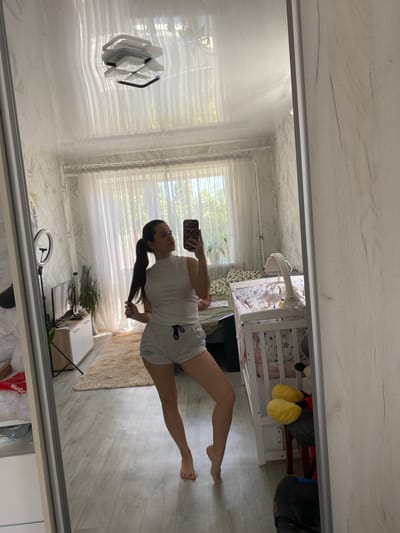 A young woman with dark hair pulled into a ponytail takes a selfie in a full-length mirror in a room. She is wearing a white sleeveless mock-neck top and gray shorts. Her left hand holds her phone, and her right hand is on her ponytail. She is standing with her legs crossed at the ankles.

The room appears to be a bedroom. To the left of the mirror, a ring light is visible, along with a television and a cabinet. A plant sits on top of the cabinet. In the background, a large window with sheer curtains is visible, and beyond that, greenery can be seen. To the right of the woman, a white crib with a mobile is present. In front of the crib, a plush Mickey Mouse toy is visible. The floor is light-colored wood laminate, and a beige rug is in the center of the room. The ceiling is white and appears to be a stretch ceiling with a light fixture. The wallpaper on the walls has a light, abstract pattern.

The image was taken in the city of Romny, Ukraine.