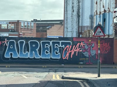 The image depicts an urban street scene in Preston, United Kingdom, under an overcast sky.
In the foreground, an asphalt road displays visible cracks, a concrete curb on the right, double yellow lines near the curb, and a broken white line marking the center.
Dominating the midground is a dark wall, approximately two meters high, extensively covered with graffiti. The central graffiti piece features "ALREET" in blue gradient bubble letters, outlined in white and black, followed by "CHA?" in dripping pink script. To the right, a pink cartoon character, resembling the Pink Panther and wearing a yellow conical hat, holds a yellow spray paint can while standing next to a white spray paint can on the ground. Below this character, the tag "@KAZAL.ONE" is visible. Further right, an unreadable red tag is present on the wall. A grey metal pole, positioned on the right, supports a triangular red-bordered sign indicating two-way traffic with two black horizontal arrows; a small bird is perched atop this sign.
The background consists of various urban structures. To the left, a multi-story brick building features multiple windows, some reflecting the sky, with visible signs including "SN" and "AZ AUTOS". To the right, a tall, partially demolished or derelict building exhibits exposed light blue vertical cladding, brickwork, and a metal exhaust duct. A distant sign partially reads "Creativity Works Dw". No people are visible.
