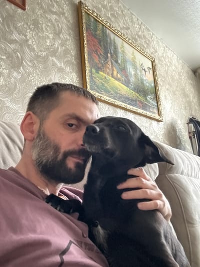 The image depicts an indoor scene in Reftinsky, Russia, featuring an adult male and a black dog. The man, positioned on the left side of the frame, has short dark hair and a well-trimmed dark beard with visible grey streaks. He wears a light purple crew-neck t-shirt, with a thin gold-colored chain visible around his neck. His right hand rests on the dog's midsection. The dog, a medium-sized canine with predominantly black fur and some grey around its muzzle, is situated to the man's right, its head tilted towards his face with eyes partially closed. Both subjects are seated on a light-colored, textured couch. The background features a wall adorned with patterned, light-toned wallpaper showcasing an ornate floral/damask design. Centered on the wall above the subjects is a rectangular, gold-framed landscape painting depicting a log cabin with a smoking chimney amidst a dense forest of evergreen and autumnal deciduous trees, with a body of water in the foreground and two dark bird silhouettes in the sky. A dark, indistinct object is partially visible on the far right of the frame, behind the couch. The scene is evenly lit.