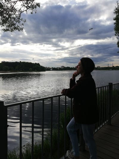 A woman stands on a wooden boardwalk, leaning on a metal railing that overlooks a large body of water. She is looking out towards the horizon, with her chin resting on her hand, appearing to be in deep thought. She is wearing a dark, possibly brown, jacket and light blue jeans. Her hair is short and dark, and she has a dark beanie on her head. The sky is overcast with dramatic, grey clouds, and shafts of light break through in places. A bird is visible flying in the distance, against the clouds. The water is rippled, reflecting the sky and the dark green trees that line the opposite bank of the lake or river. In the distance, beyond the trees, are some buildings and what appear to be industrial chimneys. The time of day appears to be late afternoon or early evening, with the diffused light suggesting an overcast sky. The overall mood is one of quiet contemplation and a connection with nature. The scene is set in Kolpino, Russia.