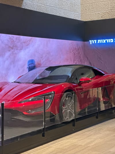 The image depicts a vibrant red sports car displayed indoors, likely in a showroom or museum. The car is positioned behind a clear barrier, possibly for protection. In the background, a large screen displays an abstract image with a sandy texture and purple fog, featuring a shadowy blue figure. Above the screen, text is visible in bright blue, appearing to be in Hebrew script, which translates to "driving miracles." The surrounding environment suggests an exhibition setting, with a textured wall above the screen and a wooden floor. The lighting is ambient, highlighting the car's glossy finish. The specific location is identified as the city of Tzafria, Israel.