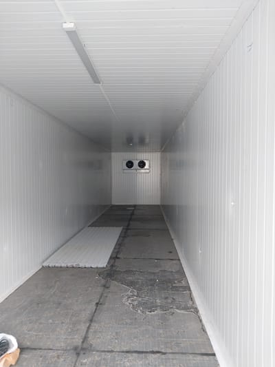 An interior view of a large, empty, white, corrugated metal storage or industrial room is shown. The room is long and narrow, with a single fluorescent light fixture mounted on the ceiling. At the far end of the room, two circular ventilation fans are visible on the wall. The floor is concrete and appears worn, with some damp or stained areas. A stack of white corrugated panels lies on the left side of the floor, near the entrance. The environment is clean and appears to be under construction or recently completed. There are no people or animals visible in the image. This scene is located in the Irgeli rural district of Kazakhstan.